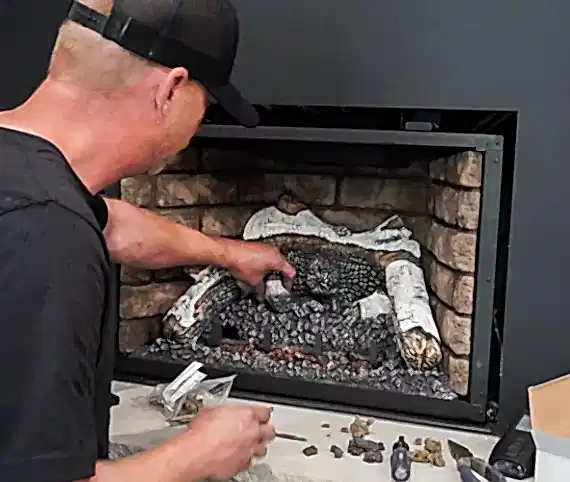 ABD technician placing ceramic logs back into a gas fireplace insert during a cleaning visit